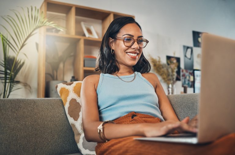 A young woman wearing glasses and a sleeveless top smiles while using a laptop on a couch in a living room decorated with plants and shelves.